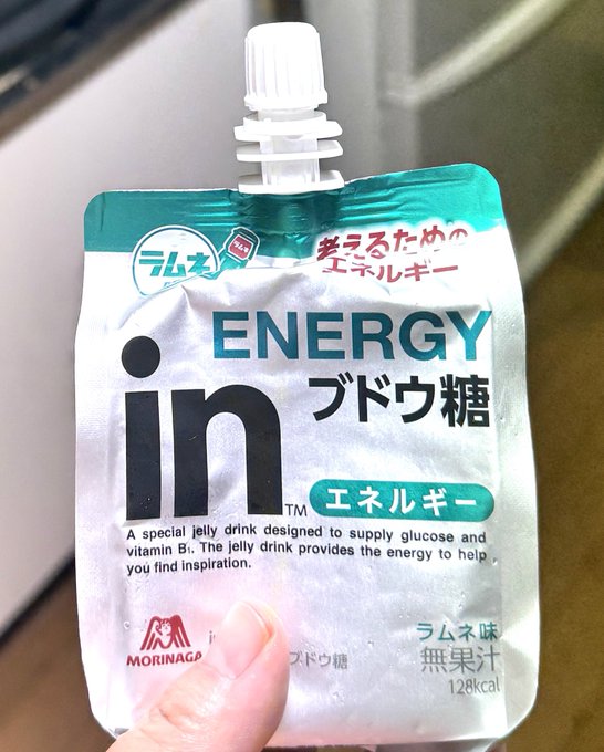 A hand holding a silver Morinaga 'in ENERGY' grape sugar (ブドウ糖) jelly drink pouch, labeled as providing glucose and vitamin B1 to fuel inspiration, with 128 kcal.