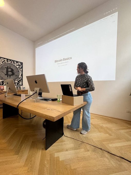A woman stands beside a wooden desk with an iMac, presenting a slide titled 'Bitcoin Basics' projected on a wall next to a Bitcoin tapestry.