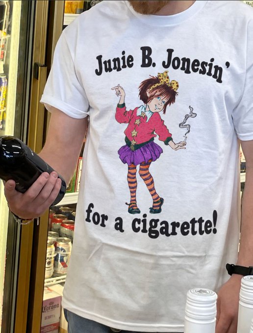 A man wearing a white t-shirt with a cartoon girl graphic and the text 'Junie B. Jonesin' for a cigarette!', holding a black bottle in what appears to be a convenience store.
