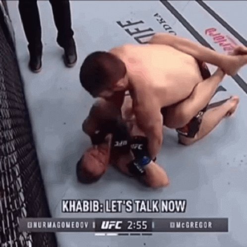 The image shows a man wearing a black shirt being pinned down by another man during a fight, with the man who is pinning him down wearing a black shirt as well. The fighter on top of his opponent appears to be in a position where he could potentially knock out his opponent. The fighters are both wearing gloves and there's a referee visible behind them, possibly observing the match or providing guidance.