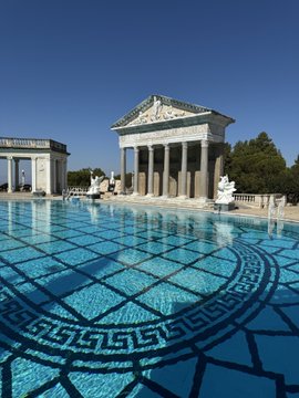 A classical-style temple with columns and a triangular pediment sits at the edge of a large blue pool with a decorative Greek key pattern on its floor.
