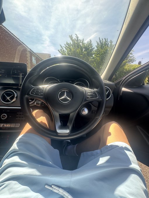 A driver's POV inside a Mercedes-Benz vehicle, showing hands resting in light-colored shorts behind the iconic three-pointed star steering wheel, with a dashboard display reading "DAB" and trees visible through the windshield—tying into the post's casual "Gm family drop SOL wallets" message about sharing crypto on a relaxed drive.