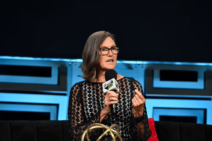 Cathie Wood, CEO of Ark Invest, speaks at what appears to be a public panel discussion while holding a microphone branded with the Ark Invest logo. The image accompanies news of her firm's recent $12.2 million purchase of SpaceX shares alongside reductions in its Block, Bullish, and Robinhood holdings.