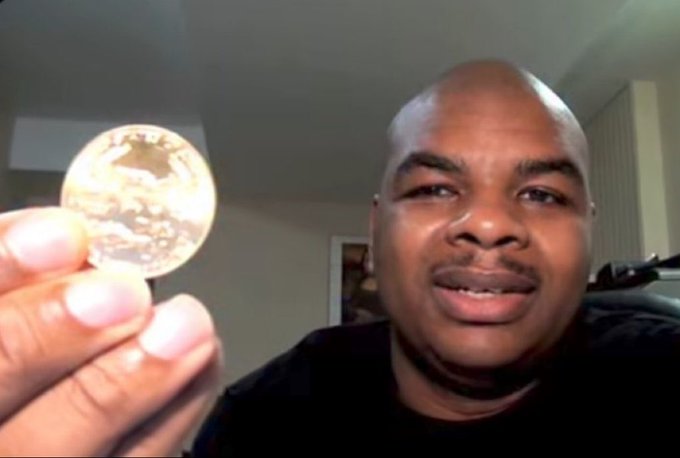 A man holds up a gold-colored commemorative coin toward the camera in what appears to be a video recording.
