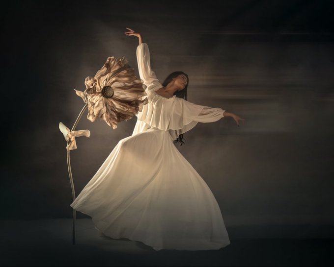 A graceful ballerina in a flowing white gown strikes a poised dance pose against a dark backdrop, her arm extended alongside an oversized golden sunflower sculpture. The image complements the post about artwork featuring flowers, capturing an elegant, ethereal floral-themed scene.