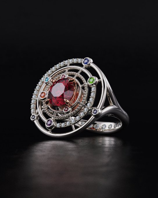 An ornate ring featuring a central red gemstone surrounded by smaller colorful gemstones (blue, green, purple) set in a silver-colored metal band with diamond accents, photographed against a dark background.