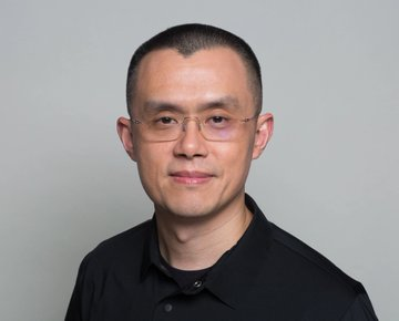 A headshot of a smiling man with short dark hair wearing glasses and a black collared shirt against a gray background.