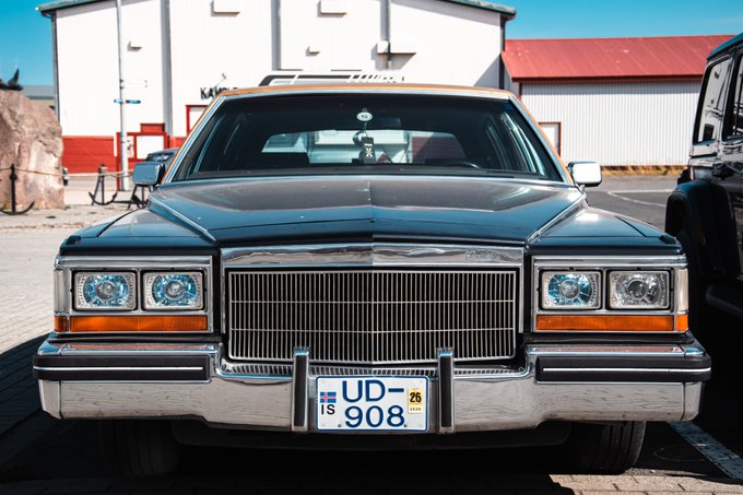 Front view of a dark teal vintage Cadillac parked outdoors, displaying an Icelandic license plate reading "UD-908" with bright blue halo headlights.