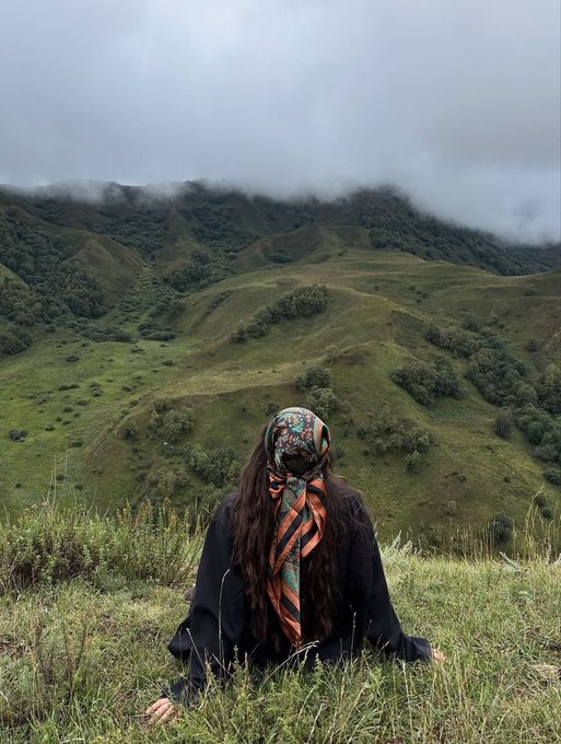 A person with long dark hair wearing a floral headscarf and black robe sits in tall grass on a misty green mountainside, facing away from the camera.