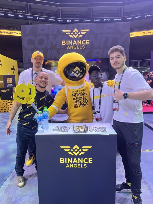 A group of five people posing at a 'BINANCE ANGELS' booth with a mascot in a yellow spacesuit at a convention event, with 'SCAN & PLAY' visible on the mascot's chest.