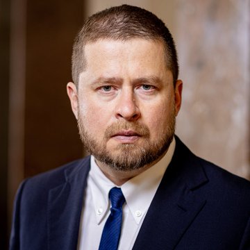 A formal portrait shows a man with short brown hair and a beard, wearing a dark navy suit, white shirt, and dark tie, looking directly at the camera with a serious expression. The image accompanies a quote from Aleš Michl discussing how adding Bitcoin to a portfolio can increase expected returns without significantly raising risk due to its low correlation with traditional assets.