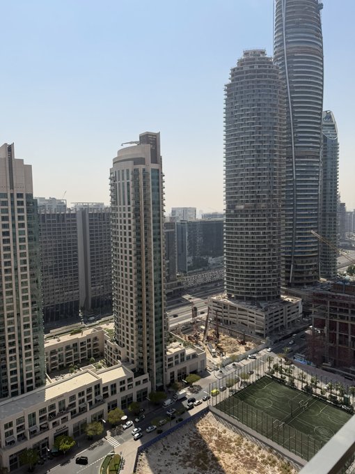 An aerial cityscape view shows tall modern skyscrapers, including a distinctive twisted tower, alongside a soccer field and roads.