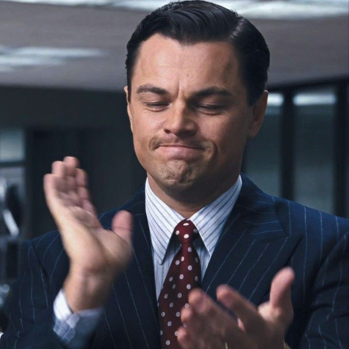 Leonardo DiCaprio in a pinstripe suit and polka dot tie clapping with a tight-lipped smile in an office setting.