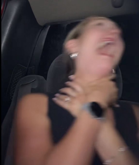 A blurry, low-quality photograph taken inside a vehicle showing a young woman laughing or screaming with her hands near her face, wearing a smartwatch and silver earrings.