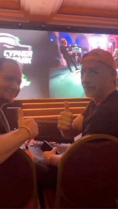 Two men give thumbs-up gestures while seated at a casino gaming table, with a screen in the background displaying 'CYPHER PUNK'.