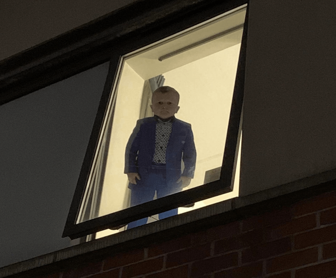 A person in a blue suit and bow tie standing in an open window at night, shot from below outside a brick building.