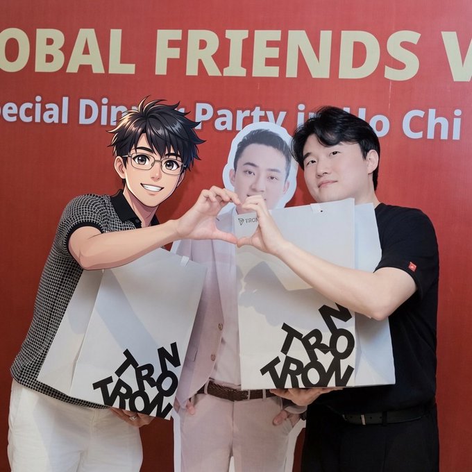 The image features two young men posing together with their hands forming a heart shape while holding white bags. The background of the photo shows a red wall with text that reads "Global Friends Vietnam" and "TRON Global Friends". This suggests that they are participating in an event or gathering related to TRON, which is a global online community for fans of anime, video games, and other forms of media. The post seems to be promoting TRON as a way to connect with fellow fans and share their passion for the series or franchise.