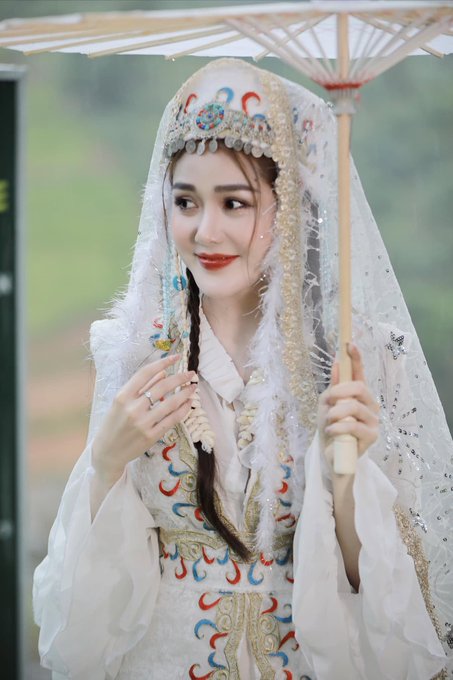A woman in elaborate, heavily embroidered traditional clothing and an ornate beaded headdress holds a parasol and smiles.