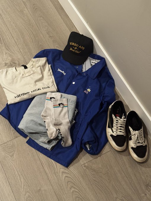 The image features a pair of shoes, a blue jacket with a black logo, and some socks lying on the floor. The shoes are white and black, while the jacket has a black and yellow logo. A hat can also be seen among these items. The socks have a cartoon character design on them. The post seems to be promoting a travel-themed collection of clothing and accessories, with a focus on casual wear for everyday use.