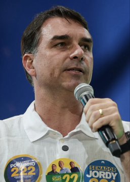 A man wearing a white shirt adorned with campaign buttons reading "BOLSONARO 22," "22," and "SENADOR JORDY" speaks into a microphone. The image relates to the post about Flávio Bolsonaro's rising odds of winning Brazil's presidency, as he is associated with the Bolsonaro political family and their right-wing movement.