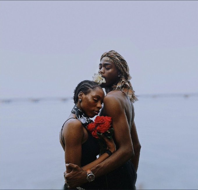 Two people embrace intimately by a misty body of water, with one holding a bouquet of red roses and wearing a patterned headscarf.