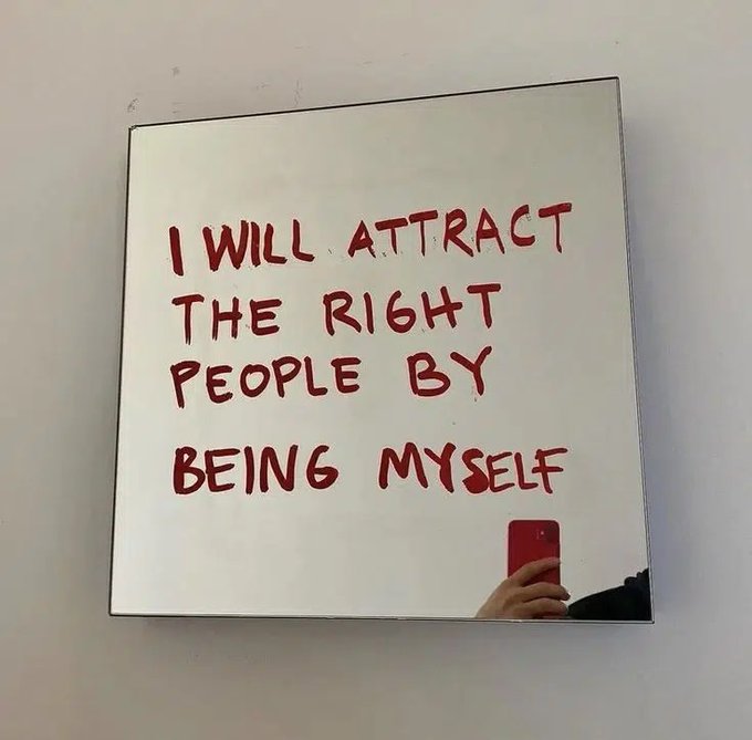 A mirror with red handwritten text reading 'I WILL ATTRACT THE RIGHT PEOPLE BY BEING MYSELF', with a person taking the photo visible in the reflection.