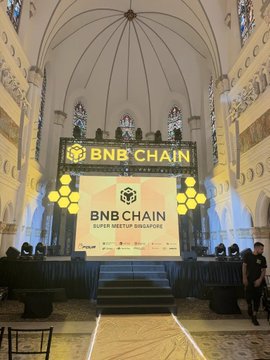 An interior view of a large hall or church featuring an ornate, high ceiling, with a stage set up for a 'BNB CHAIN' event.
