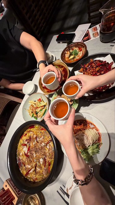 Friends clink tea cups in a celebratory toast over a lavish spread of Chinese dishes, including spicy fish, noodles, and stir-fries, marking their resilience through the bear market together.