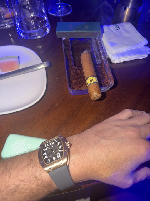A man shows off his luxury lifestyle, featuring a Cigar, a Cigar Humidor Tray, and a sleek watch (likely a Richard Mille or similar high-end timepiece), with the caption emphasizing that the rewards from trading and gambling make it all worthwhile.