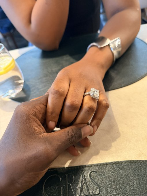 A close-up photo of two hands resting on a restaurant table, showcasing a diamond engagement ring being placed on a finger, with the brand 'GRAS' visible on the placemat.