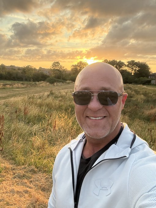 A selfie of a smiling bald man wearing sunglasses and a white Under Armour jacket, standing in a grassy field with a sunset sky and trees in the background.