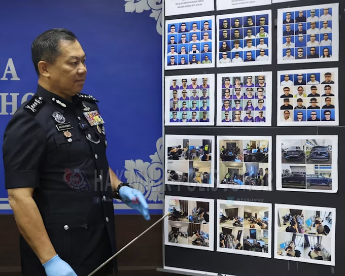 A Malaysian police officer points to a display of mugshots and evidence photos from a recent bust of telecom fraud rings in Johor, a location that is becoming a security risk. The images likely relate to the arrest of over 300 people, 95% of whom were Chinese nationals, and the investigation into a separate scam that targeted a Singaporean.