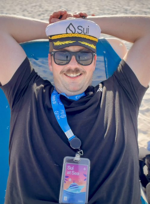 A man smiles while posing on a boat, wearing a Sui-branded captain's hat, sunglasses, and a "Sui at Sea" event lanyard, celebrating his achievement of being recognized by @SuiNetwork after 20 months of dedicated work.