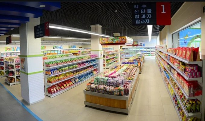 A brightly lit convenience store interior with aisles stocked with colorful packaged goods and Korean signage hanging from the ceiling, including red banners labeled '1' and '2'.