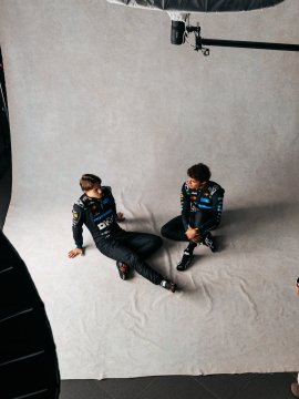 Two McLaren F1 team members in black racing suits sit back-to-back on a studio backdrop during what appears to be a behind-the-scenes photoshoot. The image relates to the post showcasing extra photos from the OKX x McLaren F1 promotional collaboration.