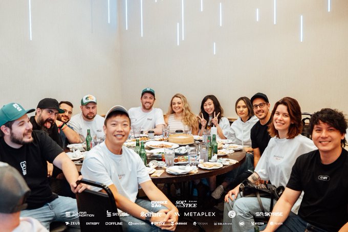 A group photo of around a dozen people gathered around a round table for a meal, with logos at the bottom including Skynet, SoftBank, Mystyle, Metaplex, Skyrise, and others.