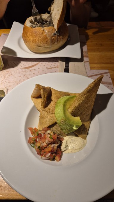 A close-up of a plated meal featuring a crepe topped with avocado slices, accompanied by a side of pico de gallo and a creamy sauce, with a bread bowl dish visible in the background.