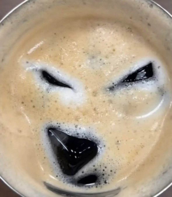 An overhead view of a creamy, frothy beverage—likely an iced coffee or root beer float—served in a metal cup, with ice cubes arranged to form an angry face expression. The dark, glossy cubes contrast against the light tan, bubbly foam, giving the drink a playful, mischievous character.