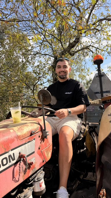A man wearing a Carhartt shirt smiles while sitting on a weathered orange tractor with "MASSEY FERGUSON" and "165" visible on the side, holding a drink with a straw. The "GM" (Good Morning) request plays on the tractor's rugged, farm-ready morning vibe — a cheerful, country-style greeting.