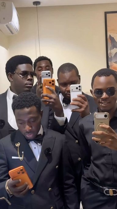 Five young men dressed in formal black suits pose closely together, with four of them holding up smartphones in various colors (orange, white, black) as if taking photos or recording.