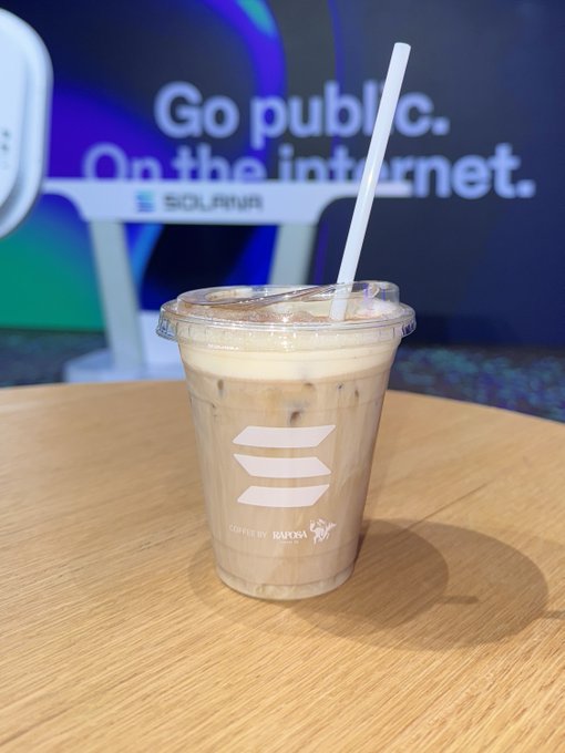 An iced coffee in a clear plastic cup with the Solana logo sits on a wooden table, with a sign reading 'Go public. On the internet.' visible in the background.