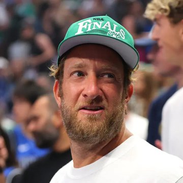 A man with a beard wearing a green "NBA FINALS" cap sits courtside with a slightly confused or exasperated expression.