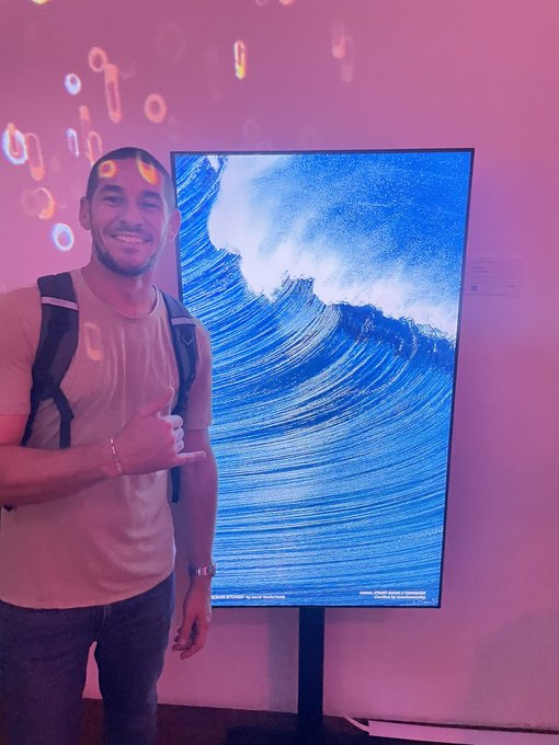 A man wearing a beige tank top and backpack posing with a 'shaka' hand sign next to a large digital screen displaying a vivid blue crashing ocean wave, with pink ambient lighting in the background.