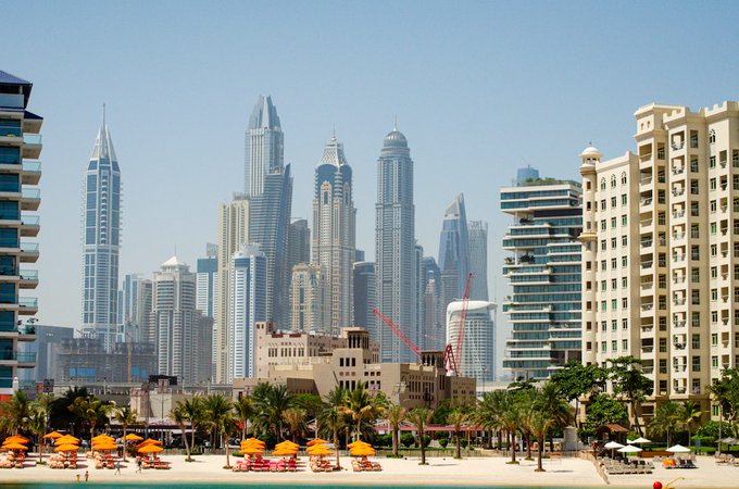 A sunny coastal cityscape showing high-rise skyscrapers and a beach with orange umbrellas and palm trees, likely depicting Dubai Marina.