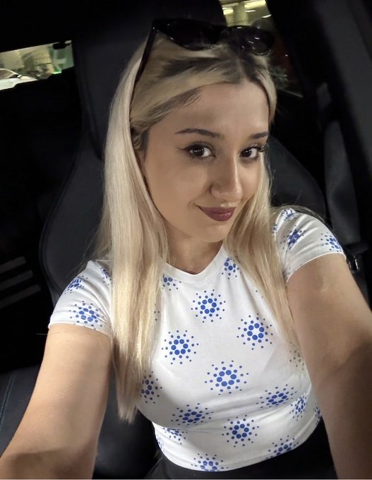A selfie of a young blonde woman wearing a white t-shirt with blue bubble patterns, sitting inside a car with sunglasses perched on her head.