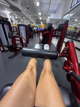 A first-person perspective photo inside a gym, showing the person's legs extended on a leg exercise machine with red equipment and other gym-goers visible in the background.
