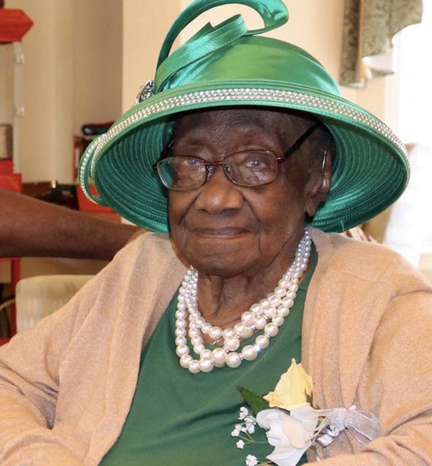 An elderly woman wearing a large green formal hat, multi-strand pearl necklace, and a cream-colored cardigan with a corsage smiles at the camera, her dignified yet playful appearance fitting the accompanying joke about her waiting for Ethereum to reach $10K as her "secret to a long life."