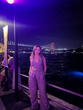 The image features a woman standing near a pier at night, wearing a purple outfit that includes a halter top and pants. She appears to be posing for a picture with her hand resting on her hip. The background of the photo shows a beautiful view of the water under a bridge in Istanbul, Turkey. The caption of the post reads "İstanbul Blockchain Week kapsamında düzenlenen #StellarParty’de Web3 topluluğuyla bir araya gelmek, yeni insanlarla tanışmak ve değerli sohbetler yapmak çok keyifliydi @StellarOrg #Web3".
