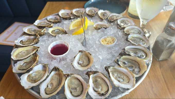 A round platter filled with crushed ice, displaying approximately two dozen fresh oysters on the half shell, garnished with lemon wedges, and accompanied by small bowls of red sauce and horseradish.