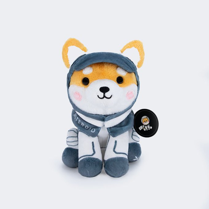 A plush toy depicting the Shiba Inu mascot 'Asteroid' dressed in a gray and white spacesuit with the name 'ASTEROID' embroidered on it.